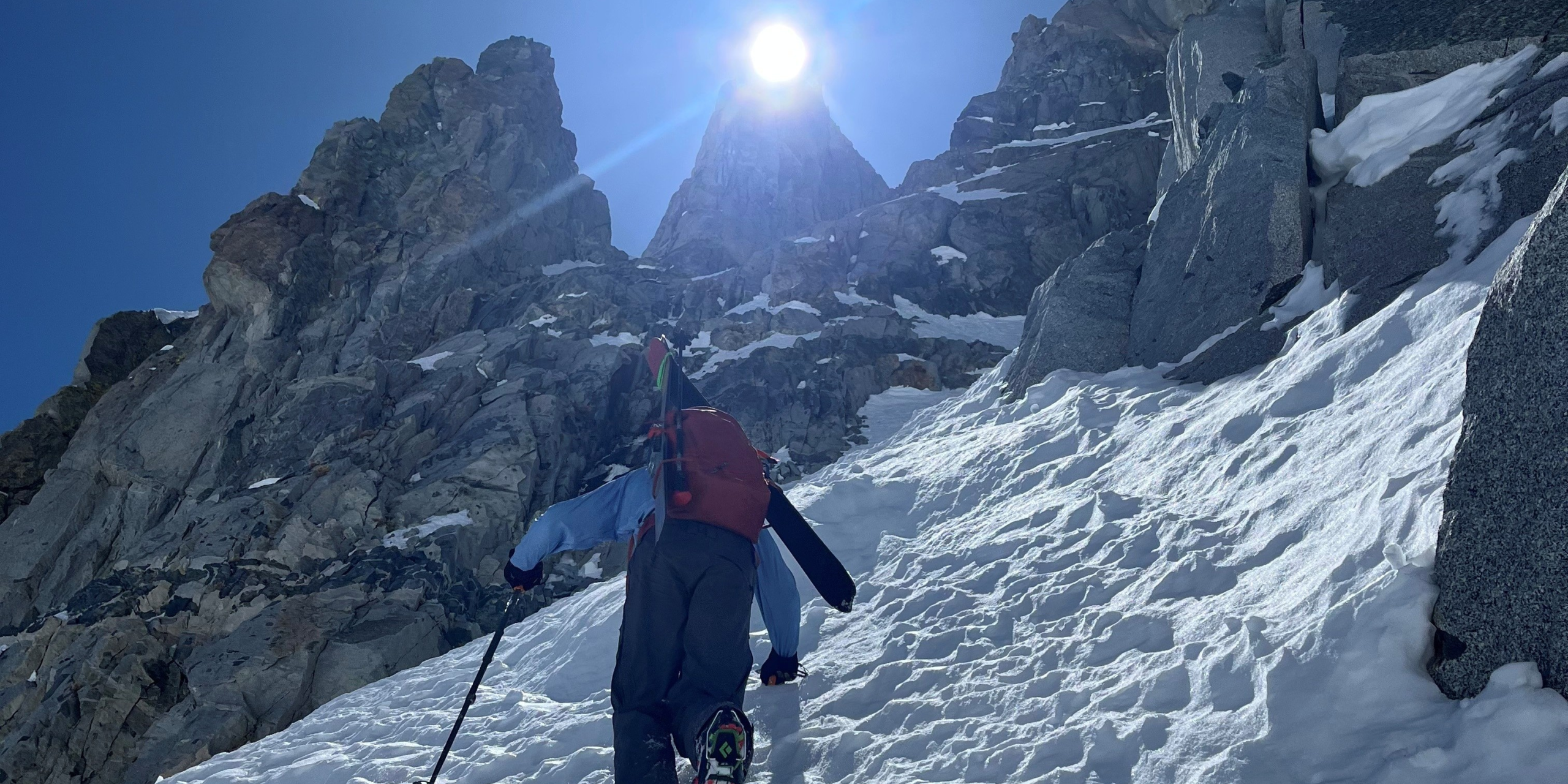 Ski mountaineer climbing on snow toward peak with sun behind it.